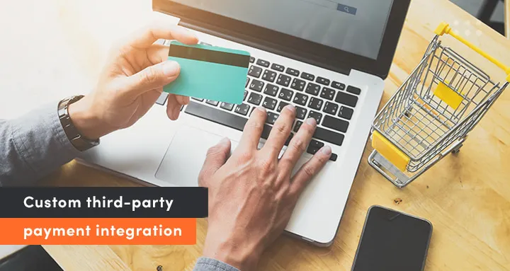 Person entering credit card details on a laptop to illustrate custom third-party payment integration in a SaaS application. A small shopping cart and smartphone are visible on the desk.