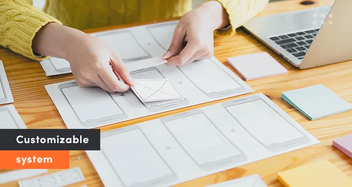 Designer arranging mobile app wireframes on a desk to illustrate customizable system design in a SaaS product. Laptop and sticky notes are visible in the background.